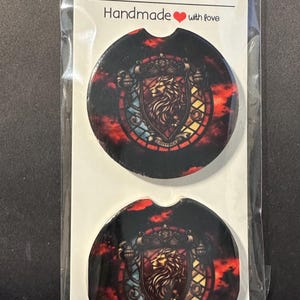 May include: Two car coasters with a stained-glass design featuring a lion crest. The coasters are black with red, yellow, and blue accents. The packaging reads "Car Coasters Handmade with love."