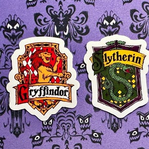 May include: Four colorful stickers featuring the crests of Hogwarts houses: Hufflepuff, Gryffindor, Slytherin, and Ravenclaw. Each crest is a unique shield design with house-specific imagery and names. The background is a purple pattern.