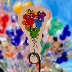 May include: A colorful sticker featuring a cluster of Mickey Mouse-shaped balloons. Each balloon is a different color, with a matching bow. The balloons are held by a cartoon hand. The background is a blurred image of balloons and a blue sky.