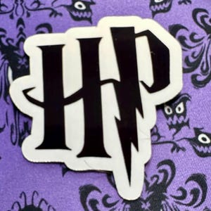 Peut inclure: Un autocollant à bordure blanche avec les initiales noires "HP" et un éclair. L'autocollant est posé sur un fond violet avec un motif répété de silhouettes noires.