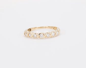 May include: A gold band ring with seven clear, round-cut gemstones. The ring has a twisted design, with each gemstone set in a bezel setting. The ring is displayed on a white background.