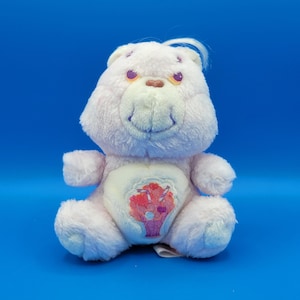 May include: A light pink Care Bear plush toy with a heart-shaped belly badge featuring a floral design in shades of pink and red. The bear has a smiling face, small ears, and a tuft of white hair. The background is a solid blue.