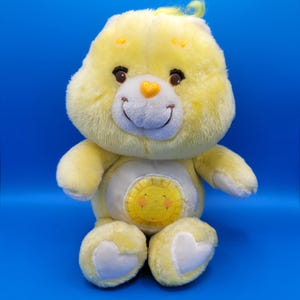 Peut inclure: Une peluche Care Bear jaune avec un visage souriant, des yeux bruns et un nez en forme de cœur. L'ours a un emblème de soleil sur le ventre et des accents blancs en forme de cœur sur les pieds. Le fond est bleu uni.