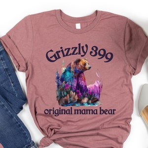 May include: Heather mauve t-shirt with a graphic of a grizzly bear in a forest setting. The text "Grizzly 399 original mama bear" is printed on the shirt.