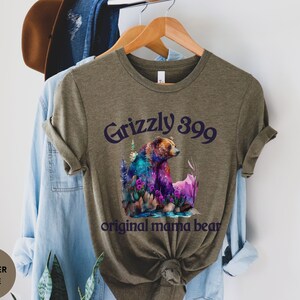 May include: A heather olive green t-shirt with a colorful illustration of a grizzly bear and the text "Grizzly 399 original mama bear".
