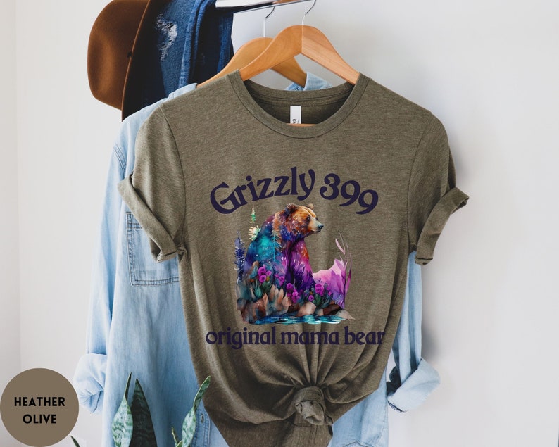 May include: A heather olive green t-shirt with a colorful illustration of a grizzly bear and the text "Grizzly 399 original mama bear".