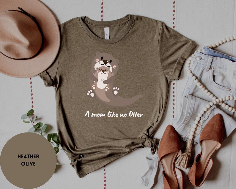 May include: A heather olive t-shirt featuring a graphic of two otters hugging. The text "A mom like no Otter" is printed below the otters.