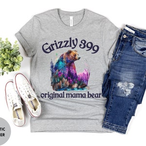 May include: Gray t-shirt with a watercolor illustration of a grizzly bear in a forest setting. The text "Grizzly 399 original mama bear" is printed on the shirt.
