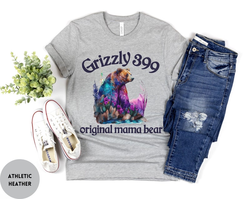 May include: Gray t-shirt with a watercolor illustration of a grizzly bear in a forest setting. The text "Grizzly 399 original mama bear" is printed on the shirt.