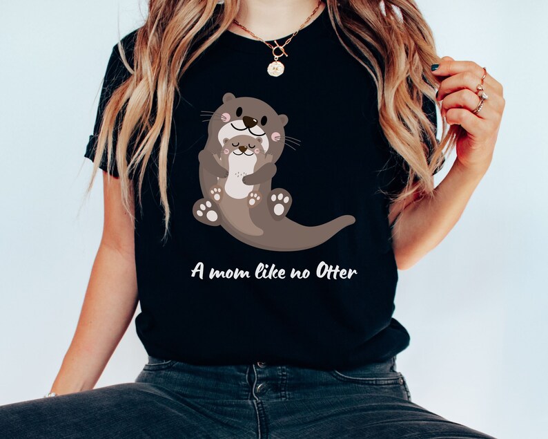 May include: A black t-shirt featuring a cartoon image of two otters hugging. The text "A mom like no Otter" is printed below the image.