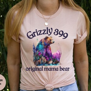 May include: A heather peach t-shirt with a watercolor illustration of a grizzly bear in a forest setting. The text "Grizzly 399 original mama bear" is printed on the shirt.