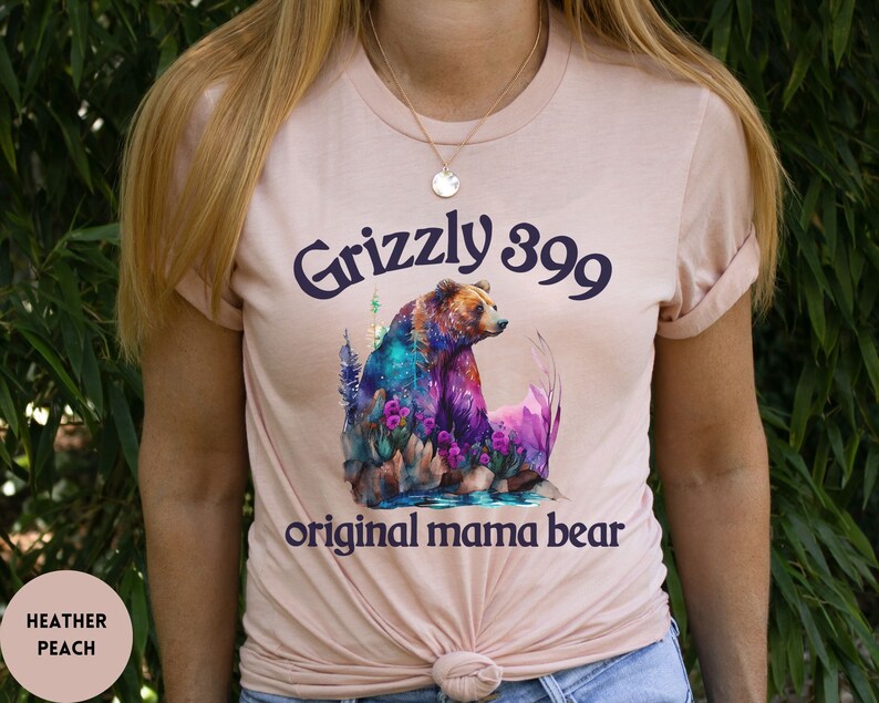 May include: A heather peach t-shirt with a watercolor illustration of a grizzly bear in a forest setting. The text "Grizzly 399 original mama bear" is printed on the shirt.