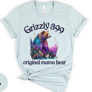 May include: Light blue t-shirt with a watercolor illustration of a grizzly bear in a forest setting. The text "Grizzly 399 original mama bear" is printed in navy blue.