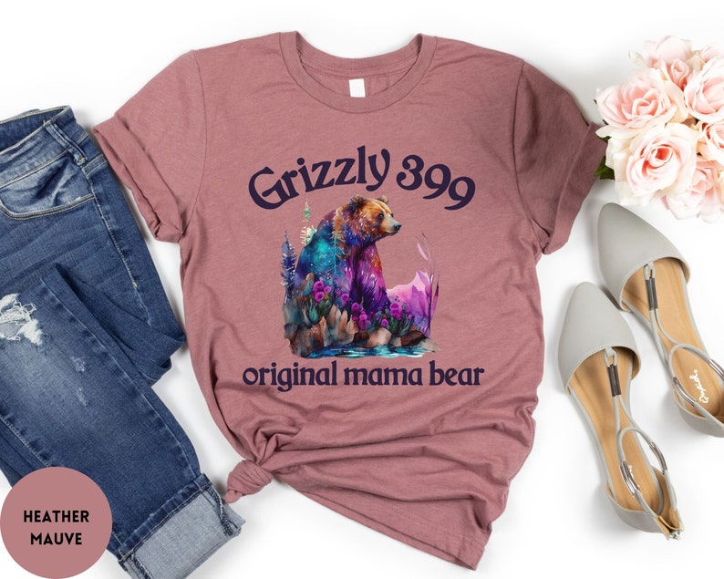 May include: Heather mauve t-shirt with a graphic of a grizzly bear in a forest setting. The text "Grizzly 399 original mama bear" is printed on the shirt.