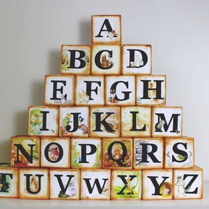 May include: A set of wooden alphabet blocks with illustrations and black letters. The blocks are stacked in a pyramid shape, spelling out the alphabet from A to Z.
