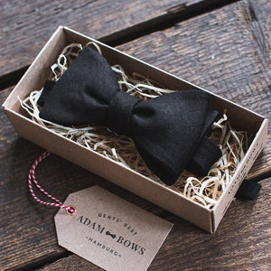 Könnte beinhalten: Ein schwarzer Fliege in einer braunen Pappbox mit zerkleinertem Papier. Die Box hat ein Etikett mit der Aufschrift "Gents' Best Adam Bows - Hamburg".