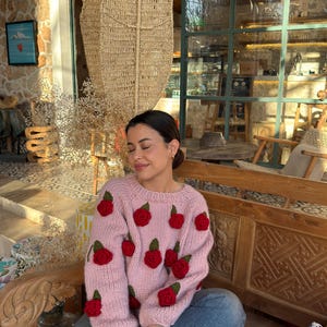 May include: A light pink sweater adorned with multiple red rose appliqués. The sweater is worn with blue jeans and tan-colored boots. The background features a wooden bench and a woven hanging decoration.