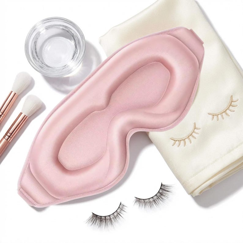May include: A light pink silk eye mask, makeup brushes, a glass of water, a white cloth with embroidered eyelashes, and a pair of false eyelashes are arranged on a white surface. The eye mask is the focal point, suggesting a beauty or self-care theme.