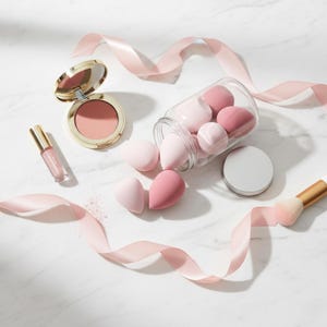 May include: A collection of makeup products, including a blush compact, lip gloss, and makeup sponges in a glass jar. A makeup brush and pink ribbon are also present. The items are arranged on a white marble surface.
