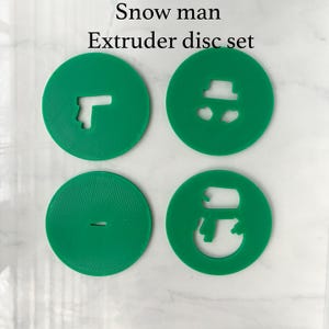 May include: A set of four circular green extruder discs with cutouts for clay or dough. The discs feature shapes including a snowman, a hat, and a small horizontal line. The text "Snow man Extruder disc set" is printed above the four green discs.