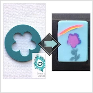 May include: A teal flower-shaped soap cutter next to a light blue soap bar with a rainbow and flower design. The soap has a pink flower and a gray stem. The soap cutter is on a white textured surface.