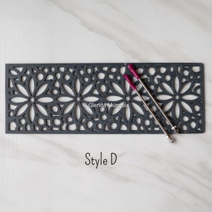 May include: A rectangular dark gray metal grate featuring a repeating floral cutout pattern. Two thin metal rods with magenta-colored tips are positioned diagonally across the surface. The text "Style D" is printed below the grate.