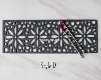 May include: A rectangular dark gray metal grate featuring a repeating floral cutout pattern. Two thin metal rods with magenta-colored tips are positioned diagonally across the surface. The text "Style D" is printed below the grate.