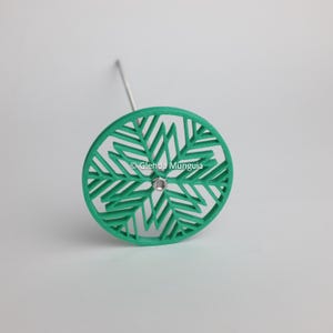 May include: A circular green object with a geometric, snowflake-like cutout pattern and a central silver metal bolt. A thin silver metal rod extends from the back of the green circular piece. The text Glenda Munguia is visible in the centre.