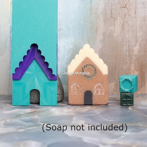 Puede incluir: Un molde de jabón en forma de casa turquesa y morada junto a un jabón marrón con forma de casa con un diseño de corona. Un pequeño cubo turquesa con un diseño de corona se encuentra encima de un cubo verde. El texto "(Jabón no incluido)" está en la parte inferior.