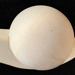 Peut inclure: Une boule en bois blanc reposant sur une base en bois blanc. La boule est lisse et ronde, tandis que la base est incurvée et légèrement allongée.