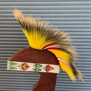 Puede incluir: Un tocado nativo americano con una pluma de color amarillo, blanco y rojo. El tocado está montado en un soporte de madera y tiene una banda de cuentas con diseños florales.