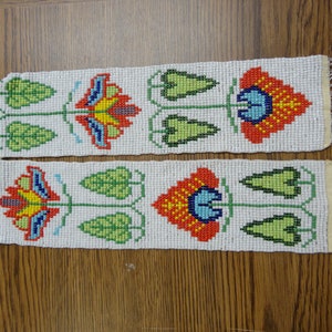 Op de afbeelding: Een kralenbies met een witte achtergrond en kleurrijke bloemenontwerpen. De bies is gemaakt van kleine kralen in tinten rood, oranje, geel, groen, blauw en wit. De bies heeft aan beide uiteinden een franje.
