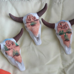 May include: Three white ceramic bull skulls with painted designs. Each skull has a different design featuring a building, cacti, and other details. The skulls are painted in shades of red, green, and brown.