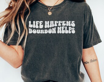 May include: A dark gray t-shirt with the text "Life Happens Bourbon Helps" in white, stylized lettering. The shirt is worn by a person with long blonde hair and a gold necklace.