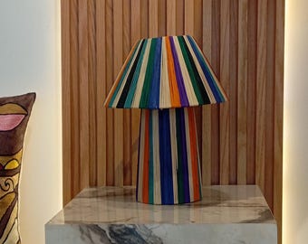 Könnte beinhalten: Eine bunte, gestreifte Lampe mit einem breiten, runden Schirm und einem hohen, zylindrischen Fuß. Die Lampe besteht aus dünnen, vertikalen Holzstreifen in verschiedenen Farben, darunter Orange, Blau, Grün und Gelb. Die Lampe steht auf einer weißen Marmorfläche.