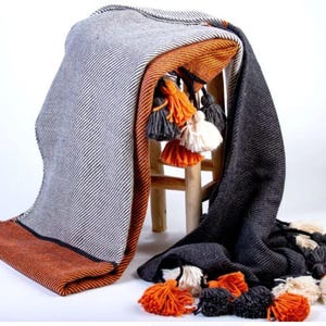 May include: A gray and orange throw blanket with a striped pattern and decorative tassels. The blanket is draped over a wooden stool, showcasing its texture and color variations. The tassels are in shades of orange, gray, and cream.