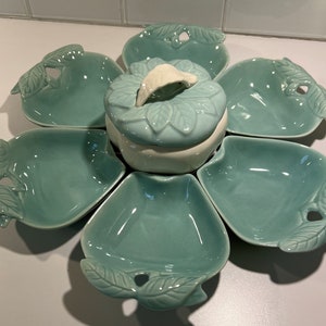 May include: A light blue ceramic serving dish shaped like a flower with six individual bowls and a central bowl with a lid. The dish has leaf-shaped handles and a white interior.