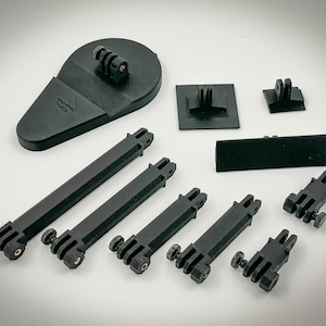 May include: A collection of black camera mounting accessories, including various brackets, mounts, and extension arms. The components are made of a matte black material and are designed for attaching cameras to different surfaces or equipment.