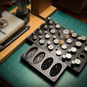 May include: A close-up of a black and silver metal press with a white base that says "Did you crease?"  The press is surrounded by a variety of small metal pieces organized in a black plastic tray.