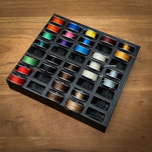 May include: A black plastic storage box with 25 compartments filled with spools of colorful thread. The spools are arranged in a grid pattern and are all different colors, including red, blue, green, yellow, and purple.