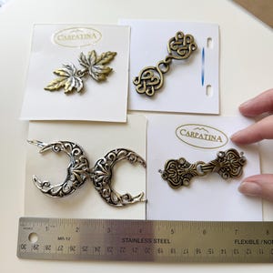 May include: Four decorative metal clasps on white cards. One is a maple leaf design, another a Celtic knot, a third is crescent moons, and the last is an ornate clasp. The cards have the brand name "CARPATINA".