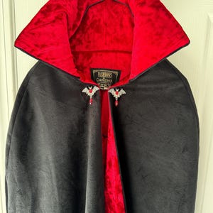 May include: Black velvet cape with a vibrant red velvet lining and collar. The cape features decorative bat-shaped clasps with red gemstone accents. The label reads "Fashions Carpatina".