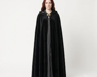 May include: A floor-length black velvet cloak featuring a hood and a gold-toned metal clasp at the neckline. The garment drapes fully from the shoulders, creating a structured silhouette that covers the torso and legs down to the floor.
