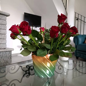 Può includere: Una vivace composizione floreale con rose rosso scuro e foglie verdi, presentata in un vaso con un design a spirale sfumato. Il vaso passa dal verde all'arancione. La composizione è su un tavolo di vetro.