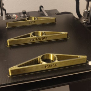 Può includere: Tre fermalibri triangolari color oro con aperture circolari e nomi personalizzati: "ELI", "ELISINA" e "VIVI". Gli oggetti sono stampati in 3D e poggiano su una superficie nera, probabilmente un piano di stampa.