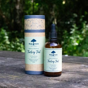 May include: A brown glass bottle with a dropper top and a label that reads "Mogo Farm Organics Medicinal Mushroom Turkey Tail 1:1 Dual Extract 30% Alcohol  Thermal Supplements Made in the UK 100ml". The bottle is sitting on a wooden surface.