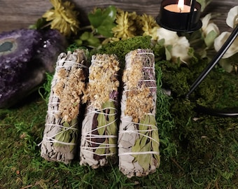 May include: Three white sage smudge sticks with dried flowers and leaves. The sticks are tied with twine and are arranged on a bed of green moss.
