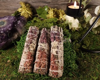 May include: Three sage bundles with dried flowers and twine are displayed on a bed of green moss. A lit candle in a black metal holder and a purple amethyst crystal are in the background. The sage bundles are a light gray color.
