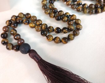 May include: A brown tassel hangs from a mala necklace made of tiger's eye beads. The necklace is strung with a dark brown cord and includes a black lava bead.