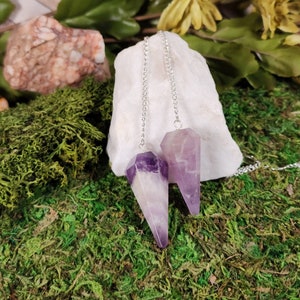 May include: Two amethyst crystal pendulums with silver chains hanging from a white rock. The pendulums are on a bed of green moss.